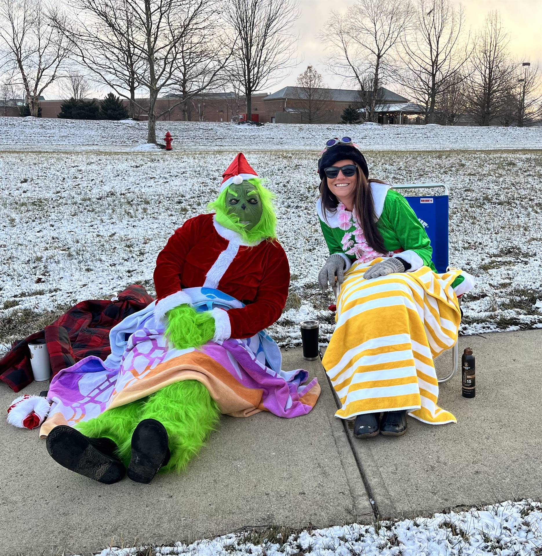 Mrs Crawford dressed as an Elf and the Grinch are sitting in beach chairs with towels greeting kids 