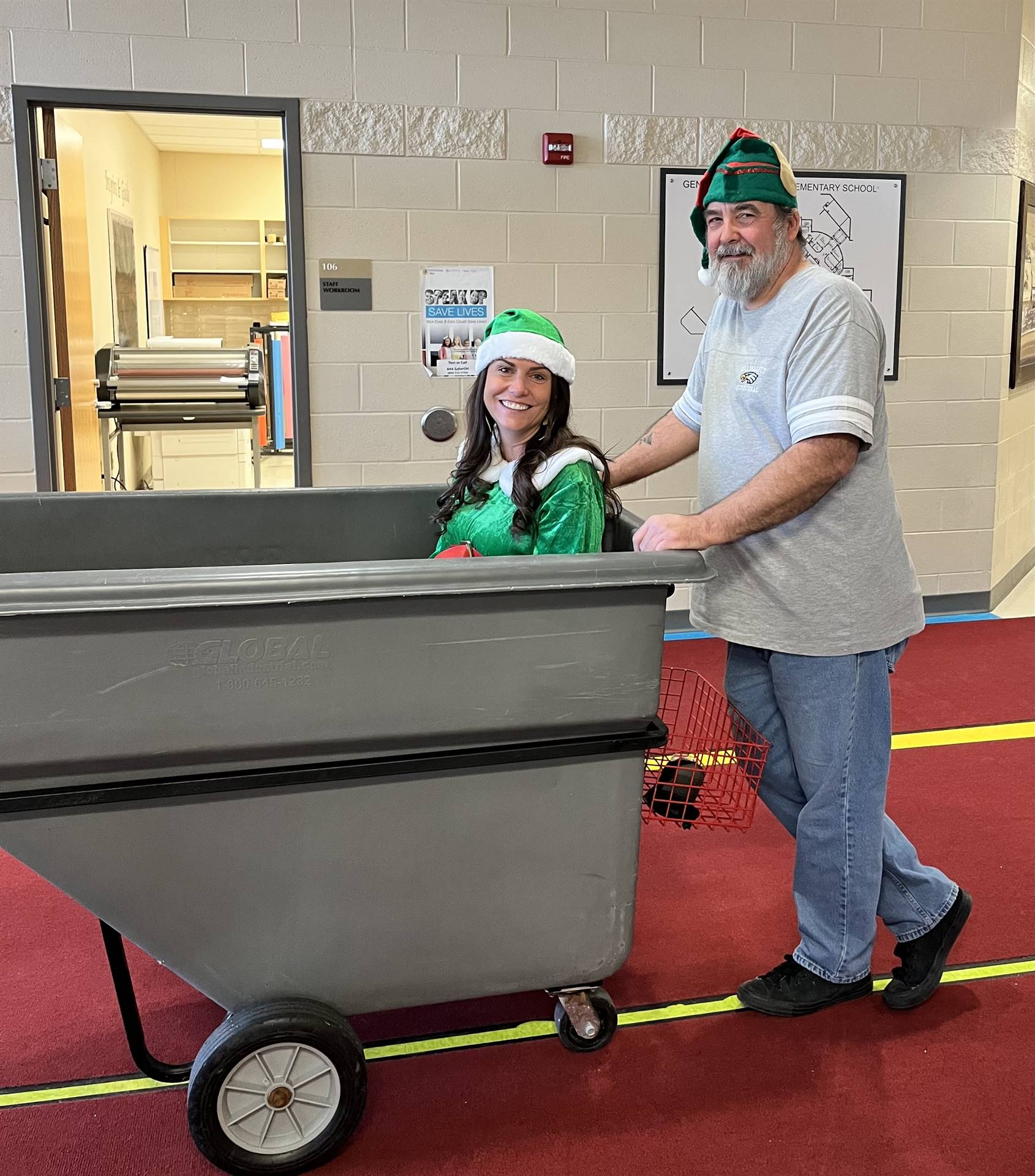 Principal dressed in Elf outfit sitting in large dumpster pushed by the custodian