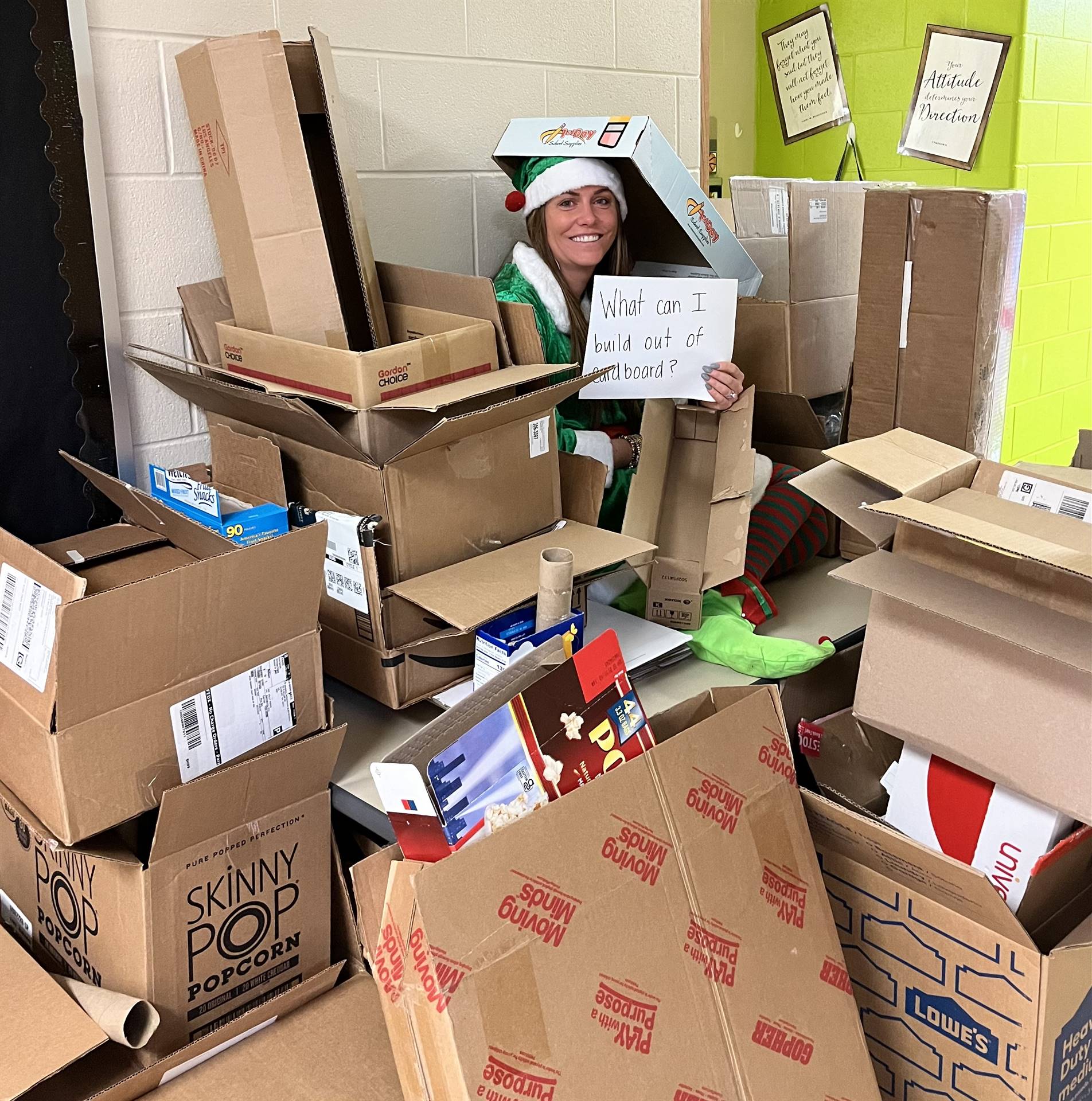 Principal Crawford is dressed in a green elf outfit sitting in a pile of cardboard. She is holding a