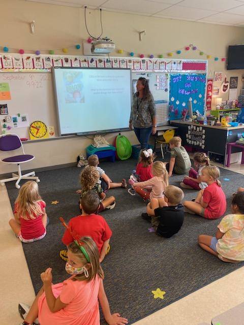 1st  grade Teacher standing at white Board while students sit on carpet listening to instruction.