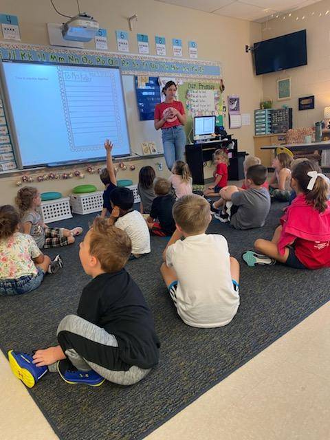 Kindergarten Teacher standing at whiteboard while students sit on carpet listening