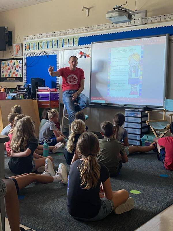 2nd Grade Teacher sitting next to Whiteboard while students sit on carpet listening to instruction