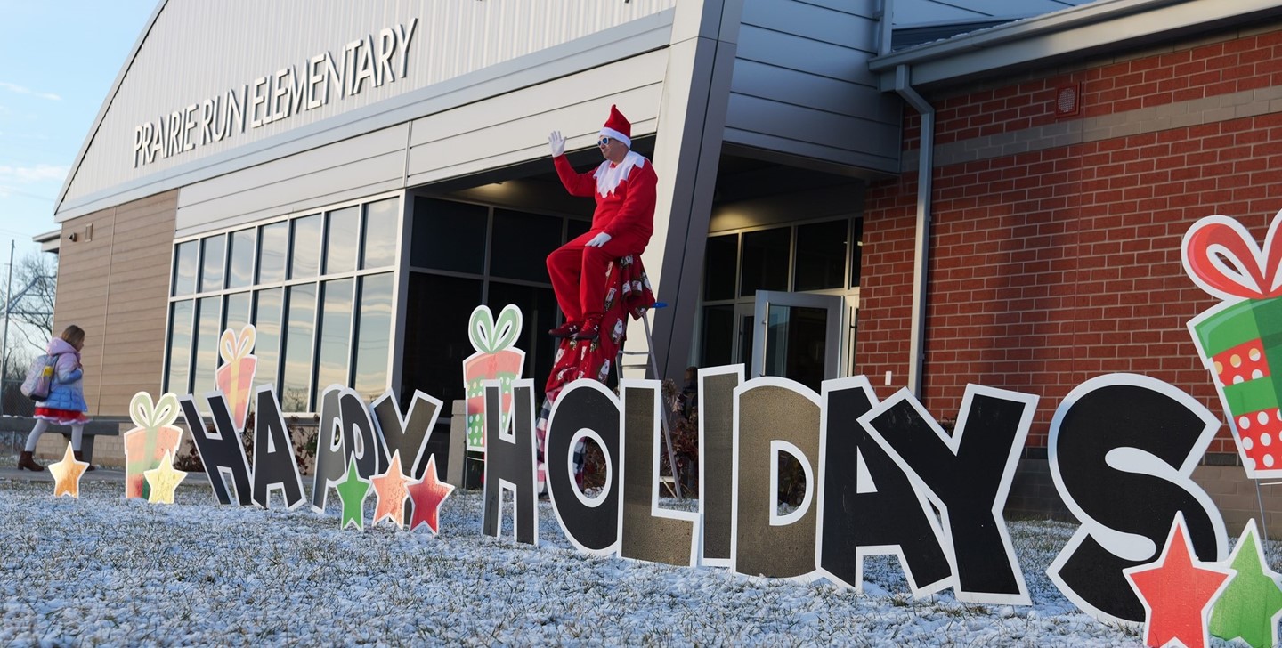 Superintendent Ryan McLane poses as an elf in front of Prairie Run Elementary - Sign reads &#34;Happy Holidays&#34;